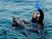 Two divers in full gear performing an emergency rescue response in open water, one diver floating on their back while the other signals for help.