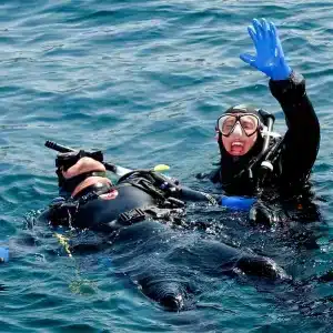 Two divers in full gear performing an emergency rescue response in open water, one diver floating on their back while the other signals for help.