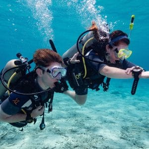 Two scuba divers underwater wearing full diving gear including masks, snorkels, and oxygen tanks, swimming side by side near the ocean floor with clear blue water surrounding them.